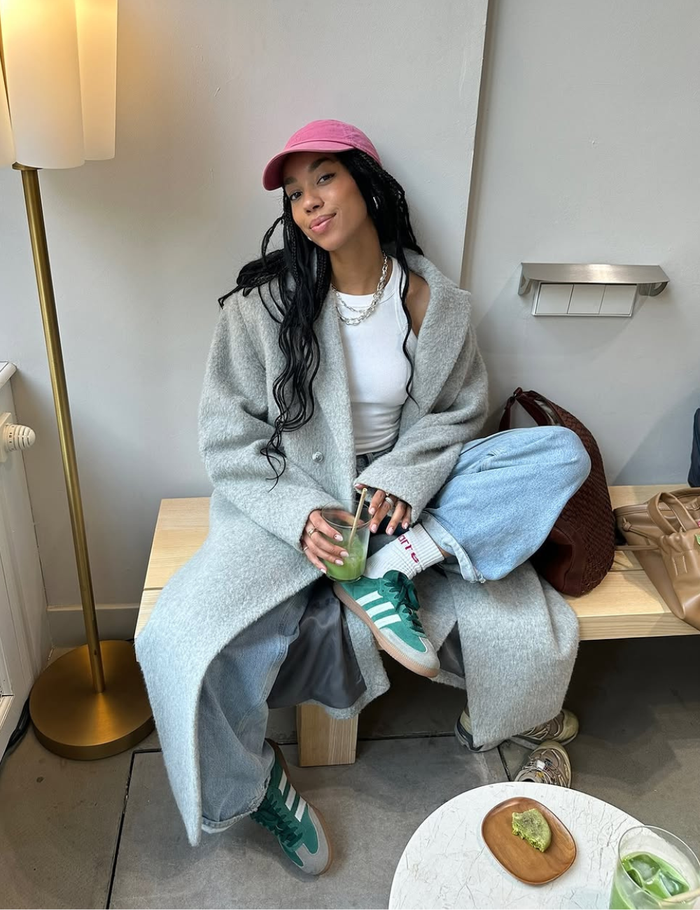 Pessoa sentada em banco, usando tênis verde e branco. Veste casaco cinza, calça jeans e boné rosa, segurando bebida verde.