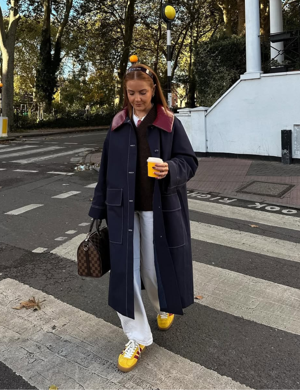 Pessoa cruzando a rua com casaco azul e calças brancas, usando tênis amarelos. Seguro de café e bolsa marrom.