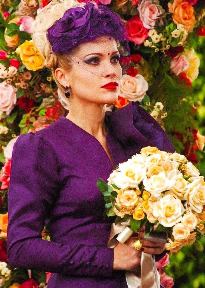 Mulher em traje elegante roxo segura buquê de rosas, com fundo colorido de flores. Elementos típicos de novelas.