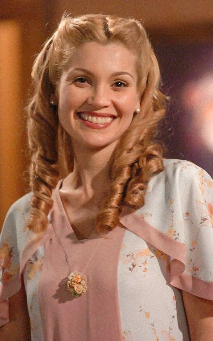 Pessoa sorridente com cabelo loiro encaracolado, vestindo blusa floral. Tema típico de novelas.