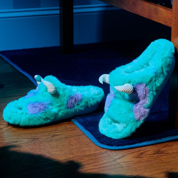Pantufas peludas verdes com chifres, inspiradas em Monstros S.A., sobre um tapete azul em piso de madeira.