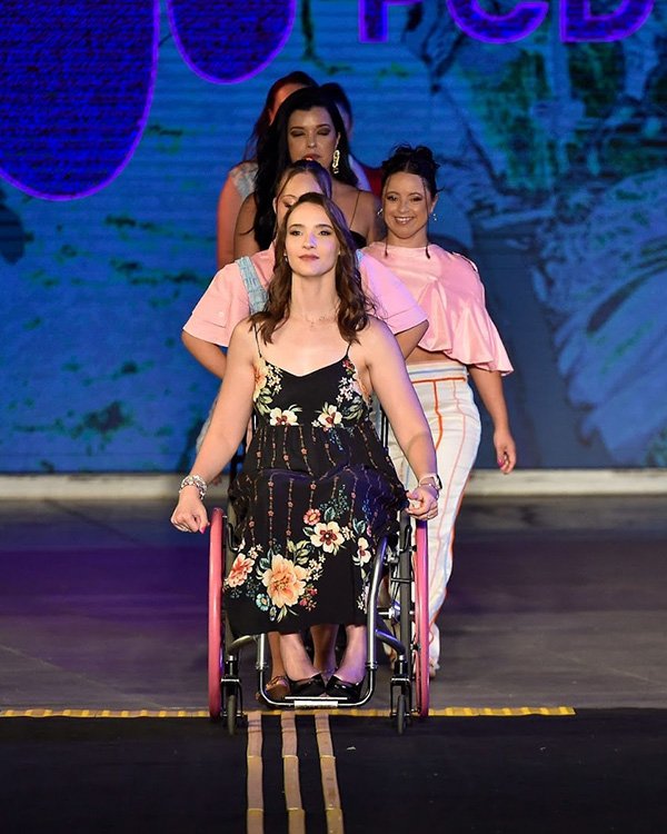 Modelo em cadeira de rodas lidera desfile de moda inclusiva, com outras modelos seguindo atrás na passarela.
