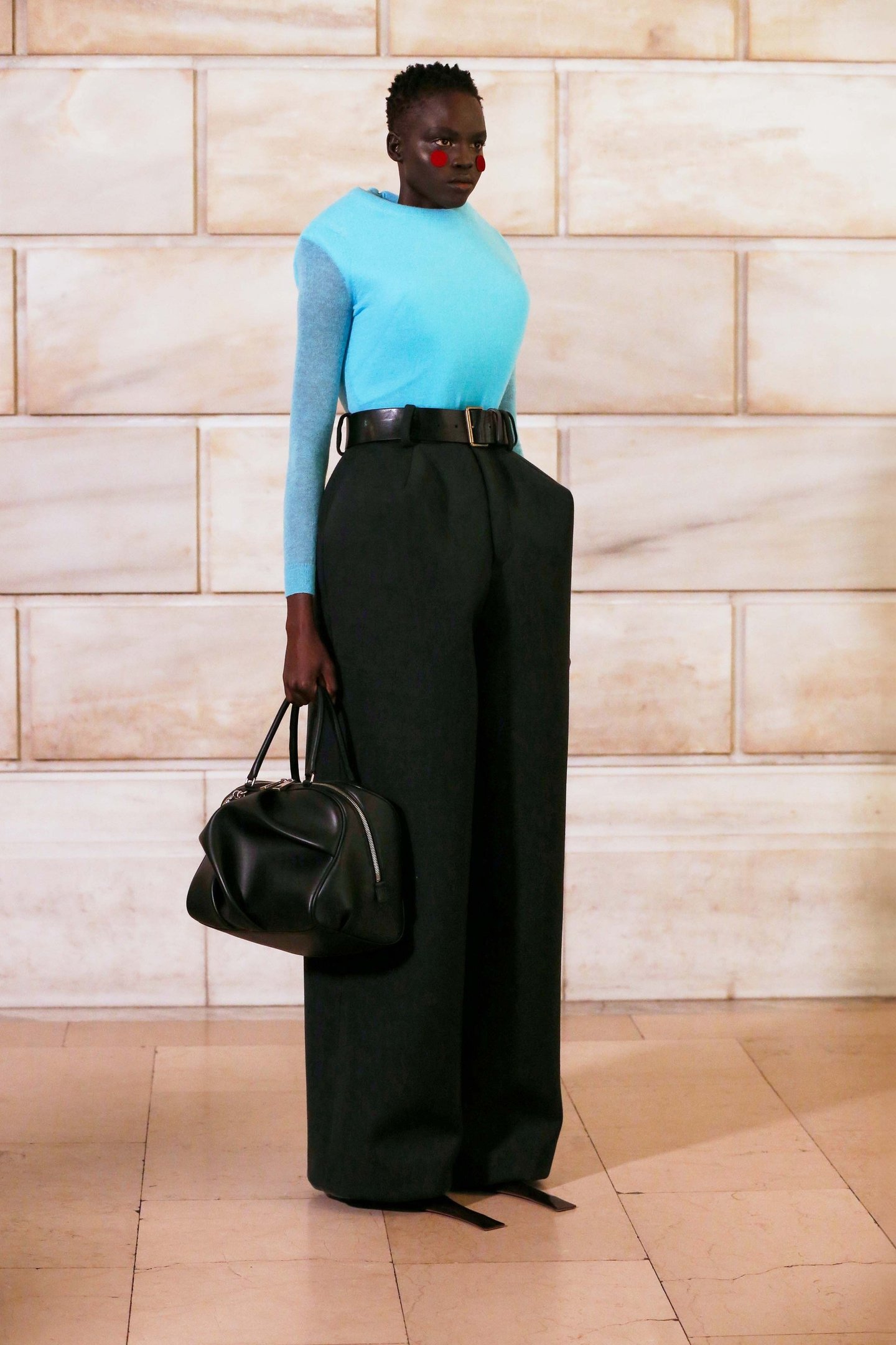 Modelo de blusa azul e calça preta, segura bolsa grande preta estilo bagagem de médico em ambiente de piso claro.