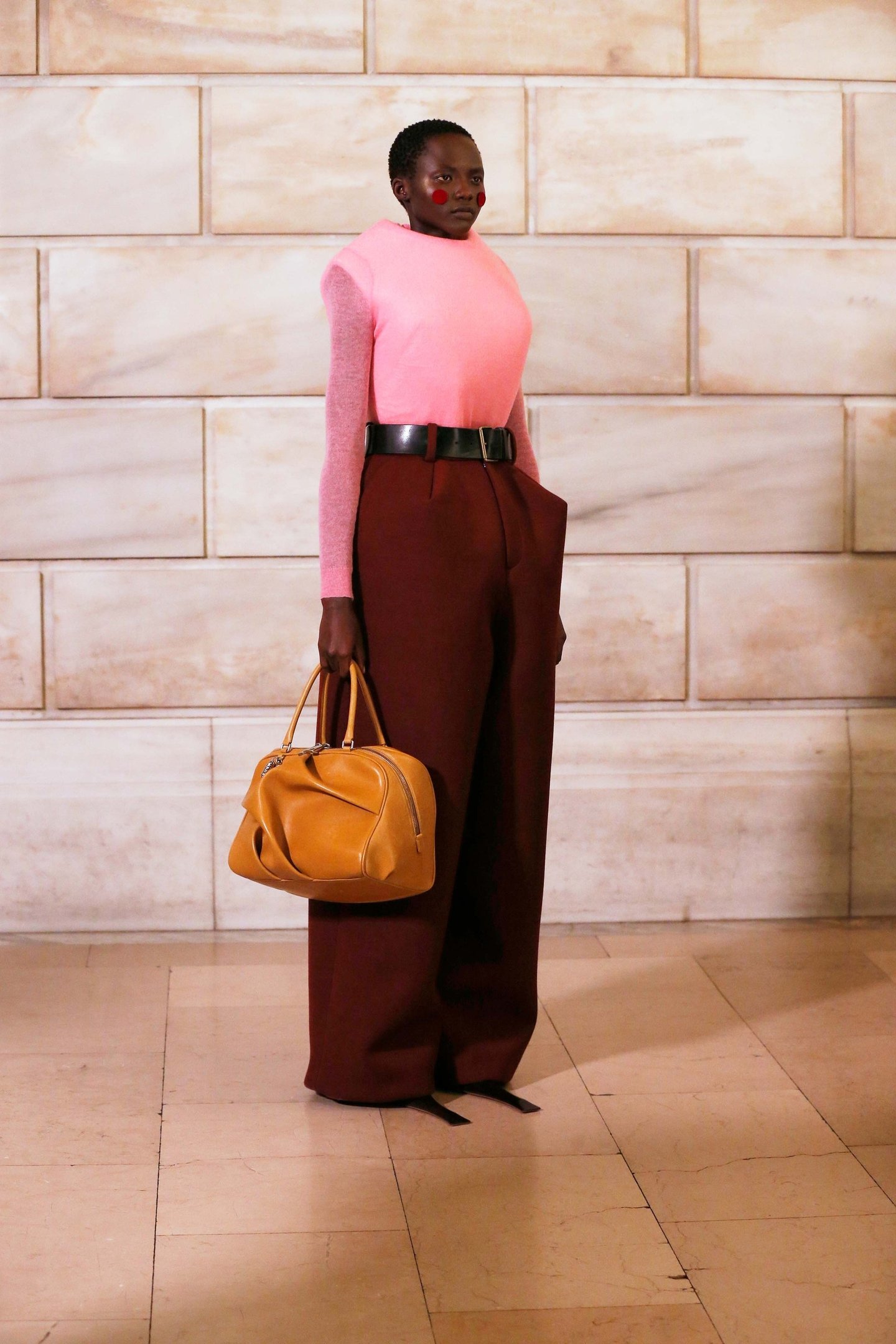 Pessoa vestida com roupa colorida segura uma bolsa estilo doctor bags marrom em um ambiente interno com parede de pedra.
