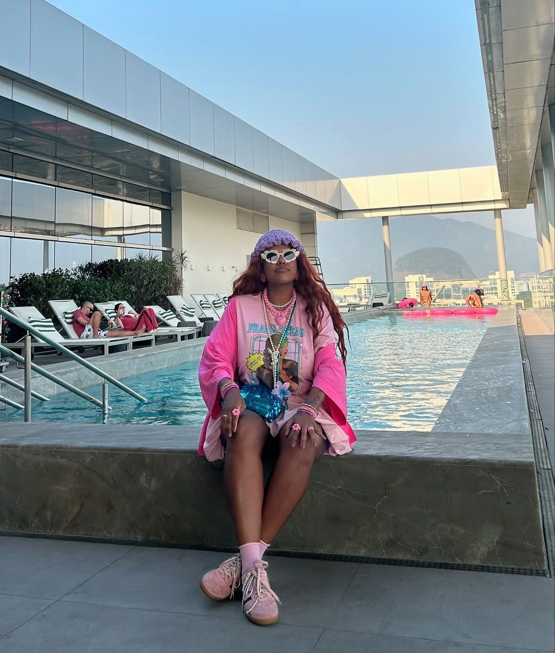Pessoa sentada à beira de uma piscina em um terraço elegante. Algumas pessoas relaxam nas espreguiçadeiras ao fundo. Ocupar espaços