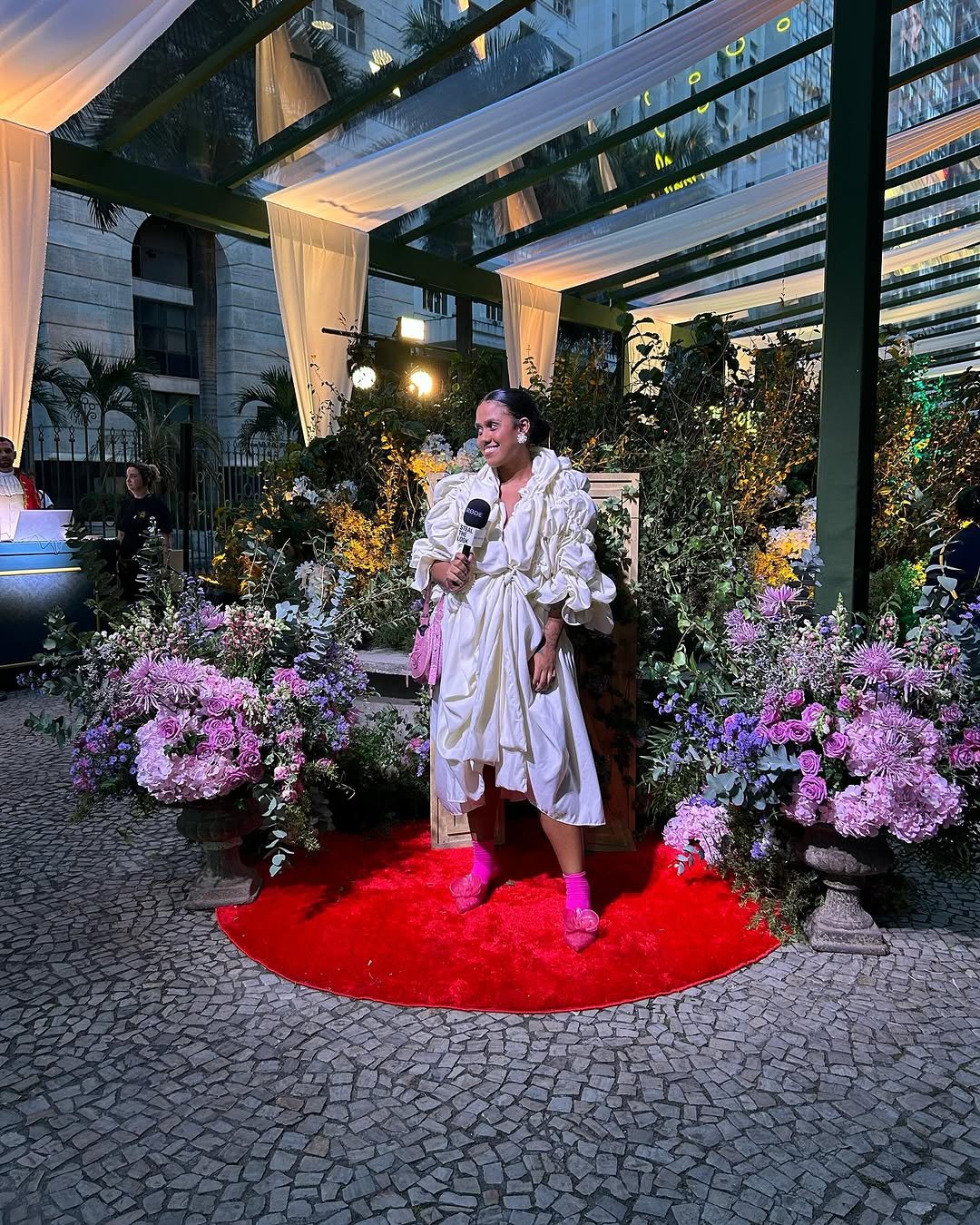 Pessoa em um vestido branco, em um evento ao ar livre decorado com flores e luzes, segura microfone. Ocupar espaços ao seu redor.