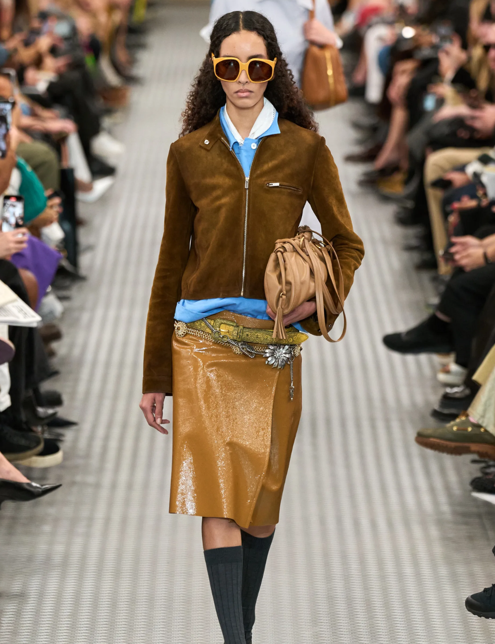 Desfile de moda com modelo usando jaqueta marrom, saia dourada e óculos de sol grandes. A palavra