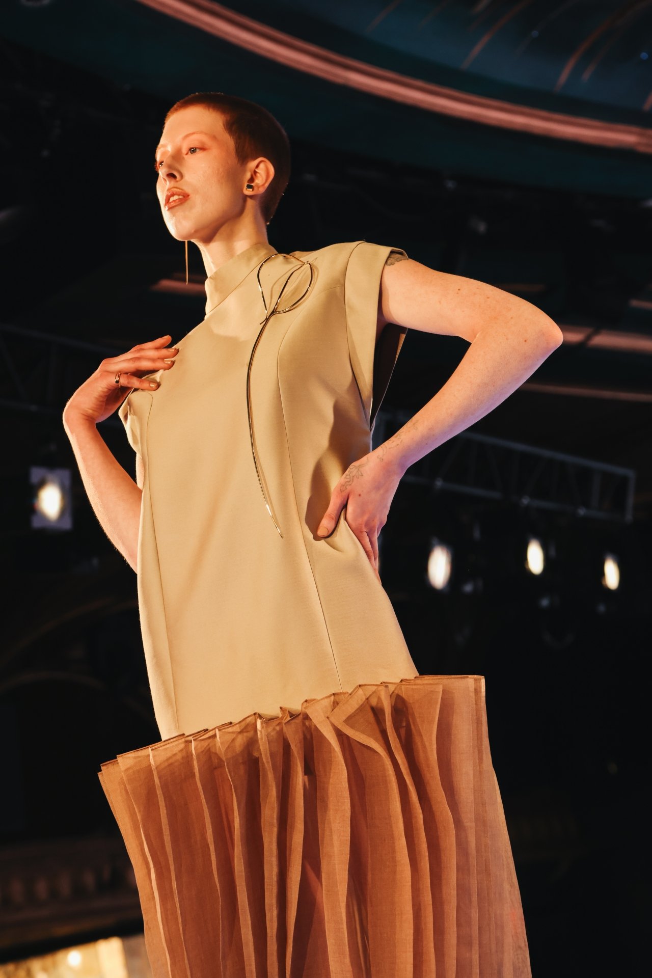 Modelo em passarela vestindo roupa elegante, em tecido bege e detalhe transparente, criando uma expressão moderna.