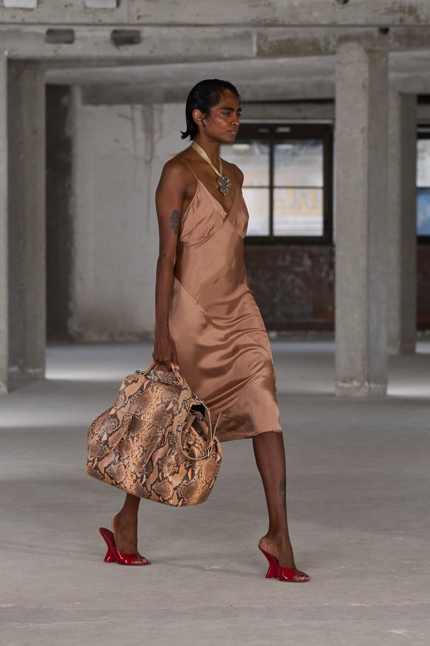 Modelo vestindo vestido acetinado marrom, bolsa com estampa de cobra e sapatos vermelhos.
