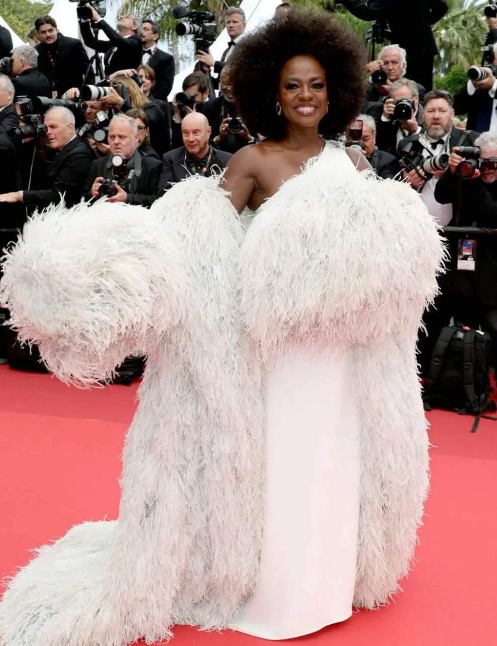 Pessoa famosa em evento, vestindo um imponente vestido branco com penas. Fotógrafos ao fundo no tapete vermelho.
