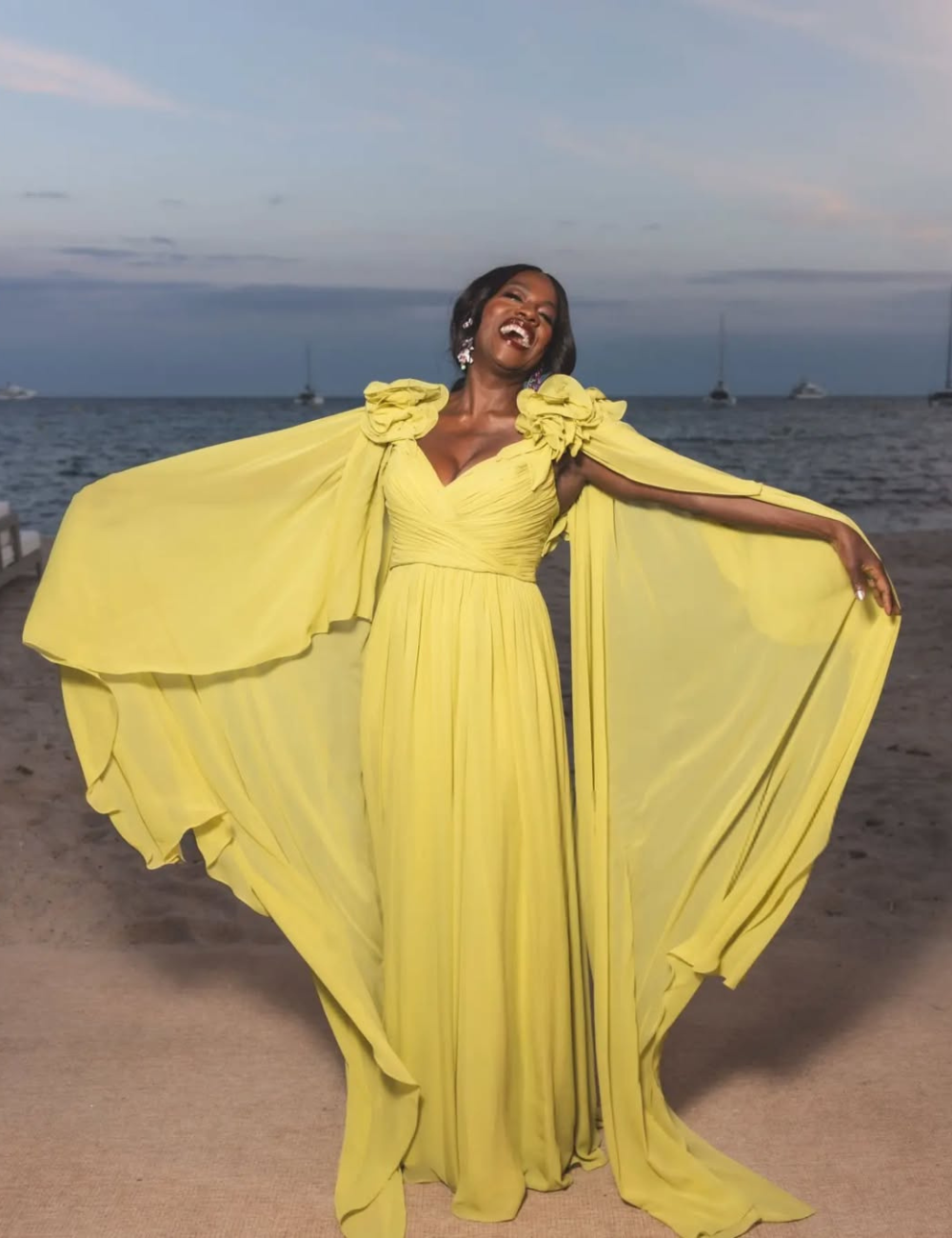 A pessoa está sorrindo, vestindo um vestido amarelo vibrante, em frente a um cenário de praia ao entardecer.