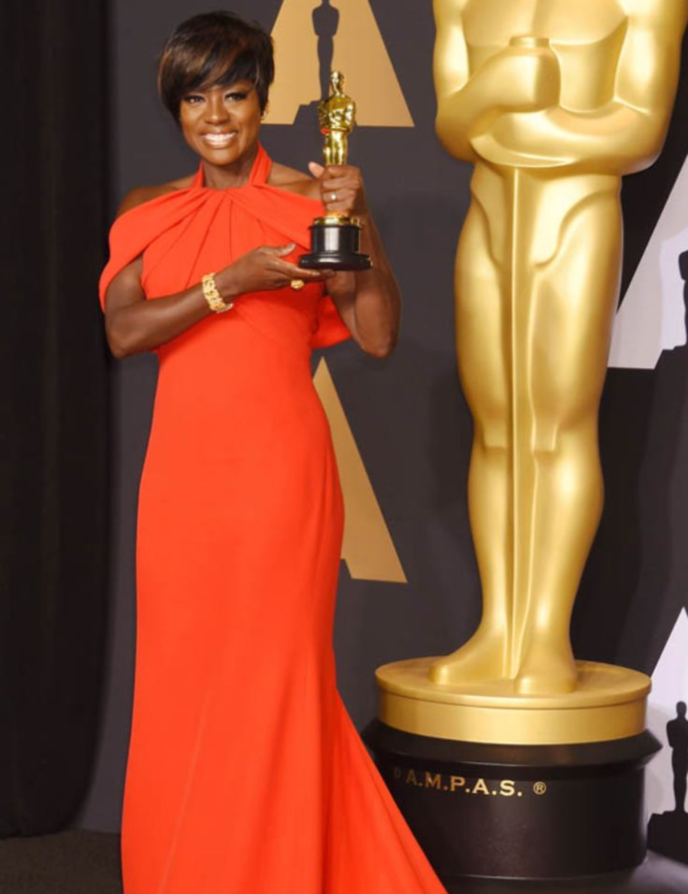 Viola Davis usando um vestido laranja, sorrindo, segurando um Oscar nas mãos.