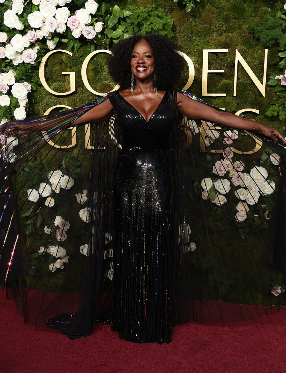 Vestido preto brilhante em evento de gala, cercado por rosas e folhagens.