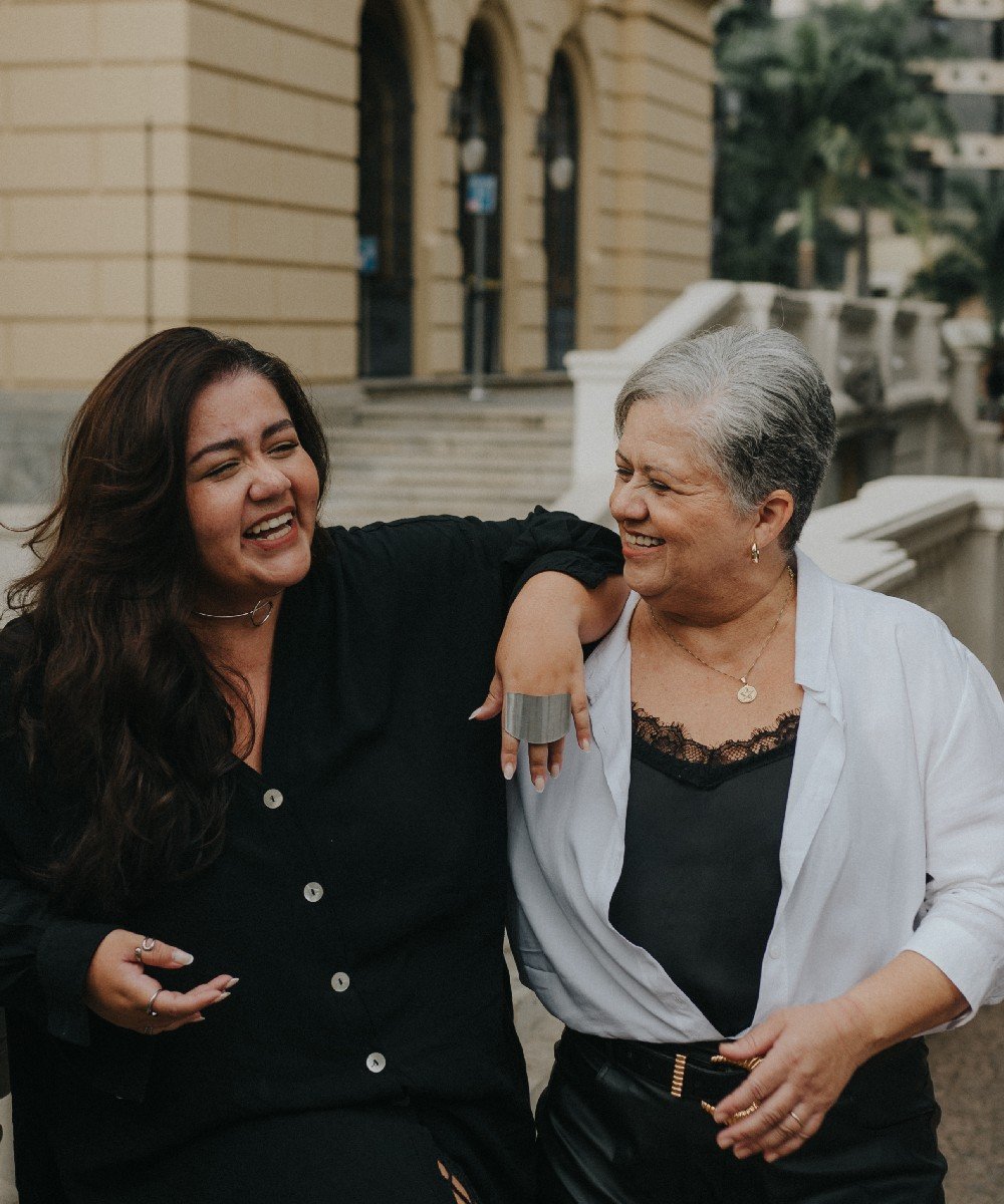 Mulheres sorrindo em frente a um prédio. A mulher à direita tem fios brancos nos cabelos.