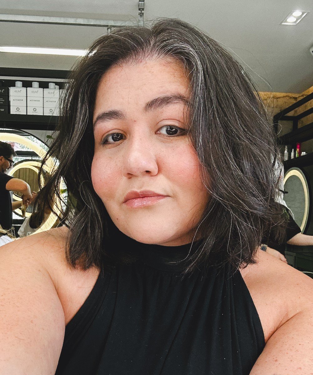 Mulher em salão de cabelo com fios brancos visíveis, veste blusa preta. Ao fundo, cabeleireiro atendendo cliente.