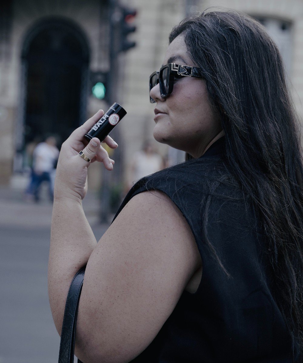 Uma mulher com fios brancos no cabelo segura um batom enquanto usa óculos escuros e veste preto.