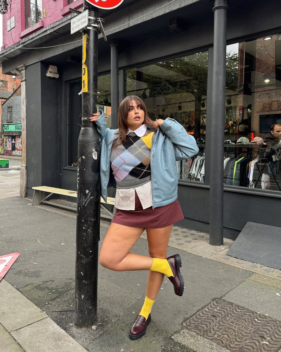 influenciadora Francesca Perks usa look colorido, saia vinho, camisa listrada, colete estampada, meias amarelas e mocassim marrom, combinando com a bolsa tendência de 2025