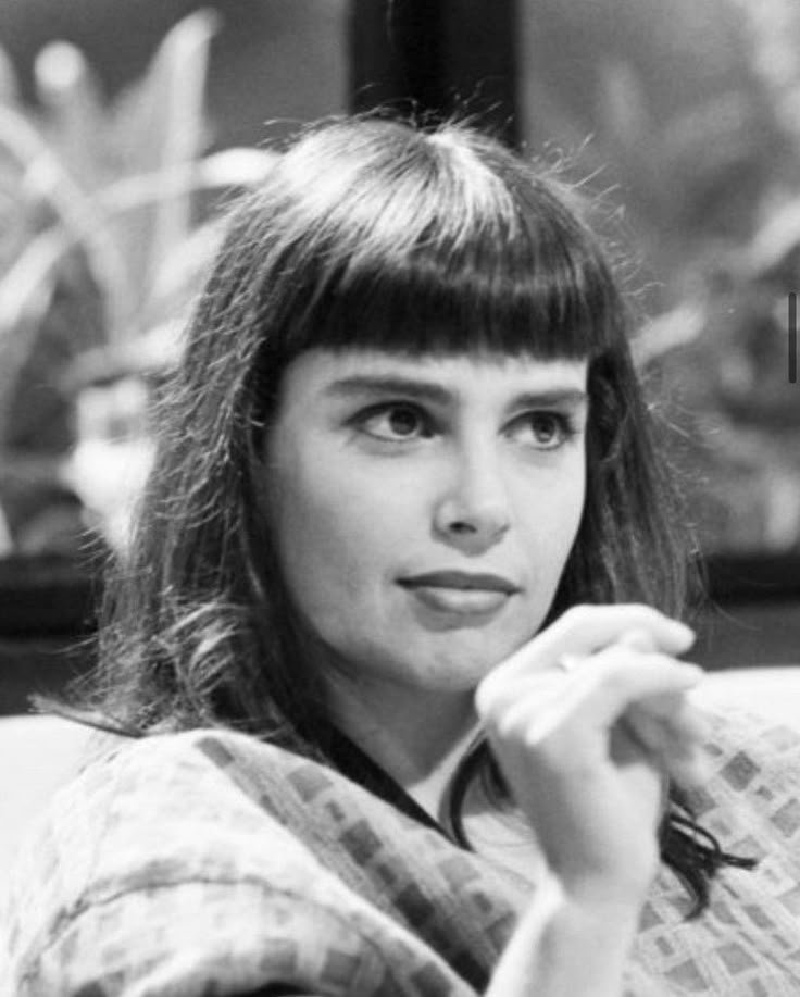 Uma mulher de cabelos escuros e franja em uma cena. Ambiente remete a um tom de Novelas.