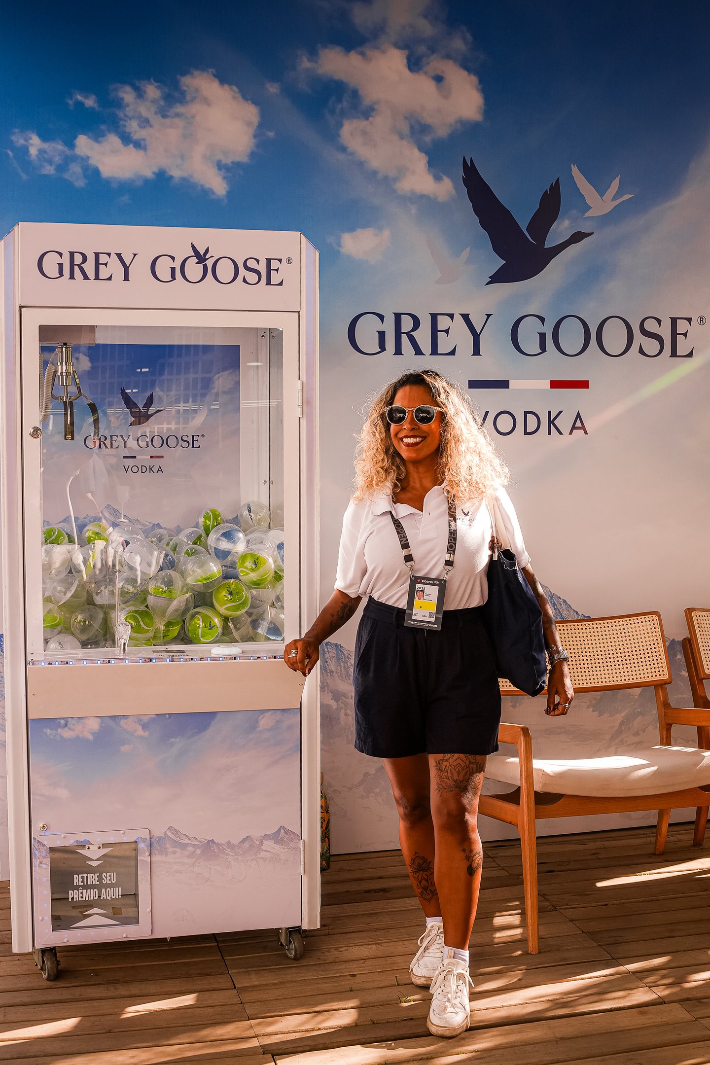 Pessoa sorrindo ao lado de uma máquina de pegar prêmios, durante o evento Rio Open. Banner Grey Goose no fundo.