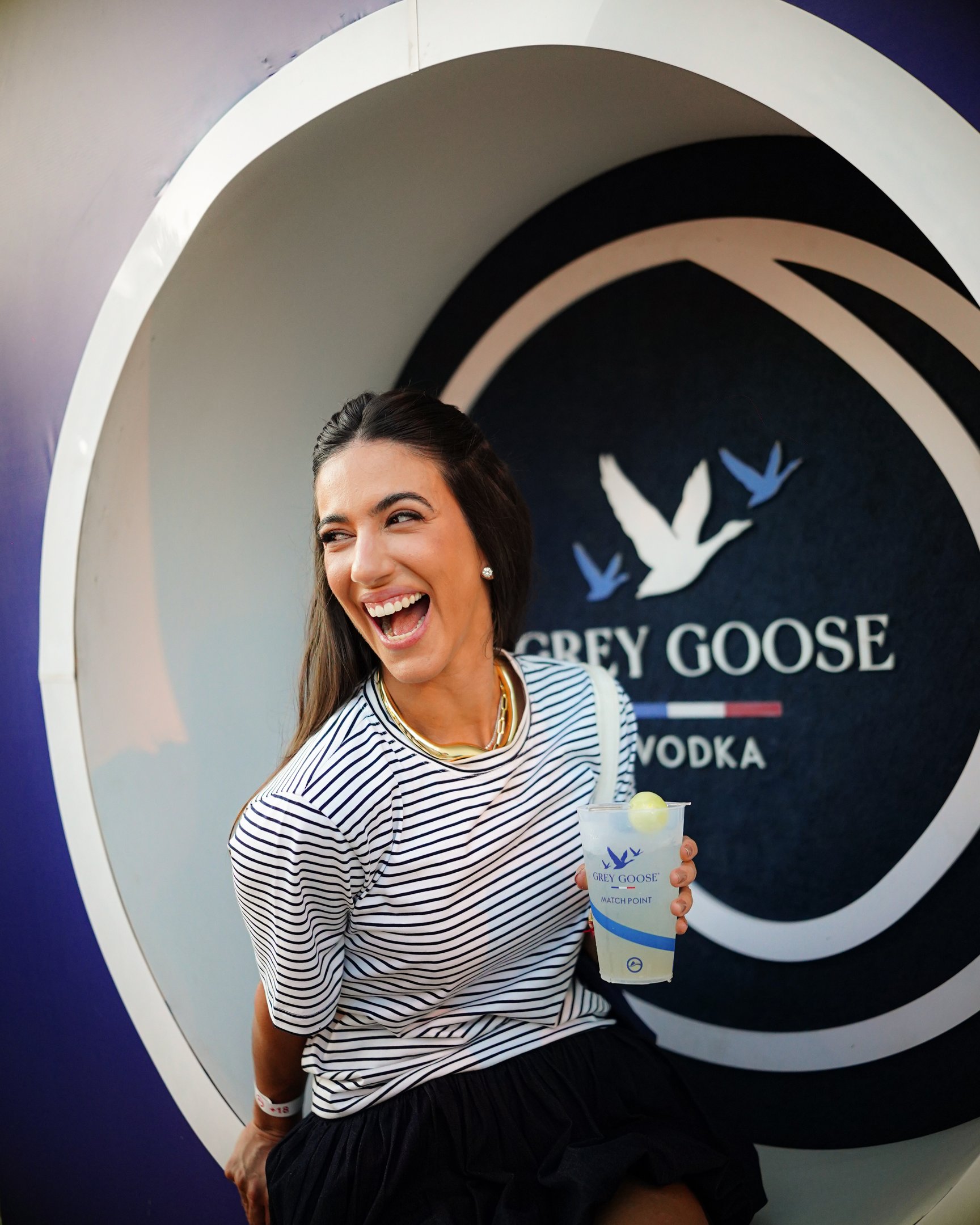 Pessoa sorrindo com copo na mão, frente a logo da Grey Goose. Ambiente descontraído no contexto do Rio Open.