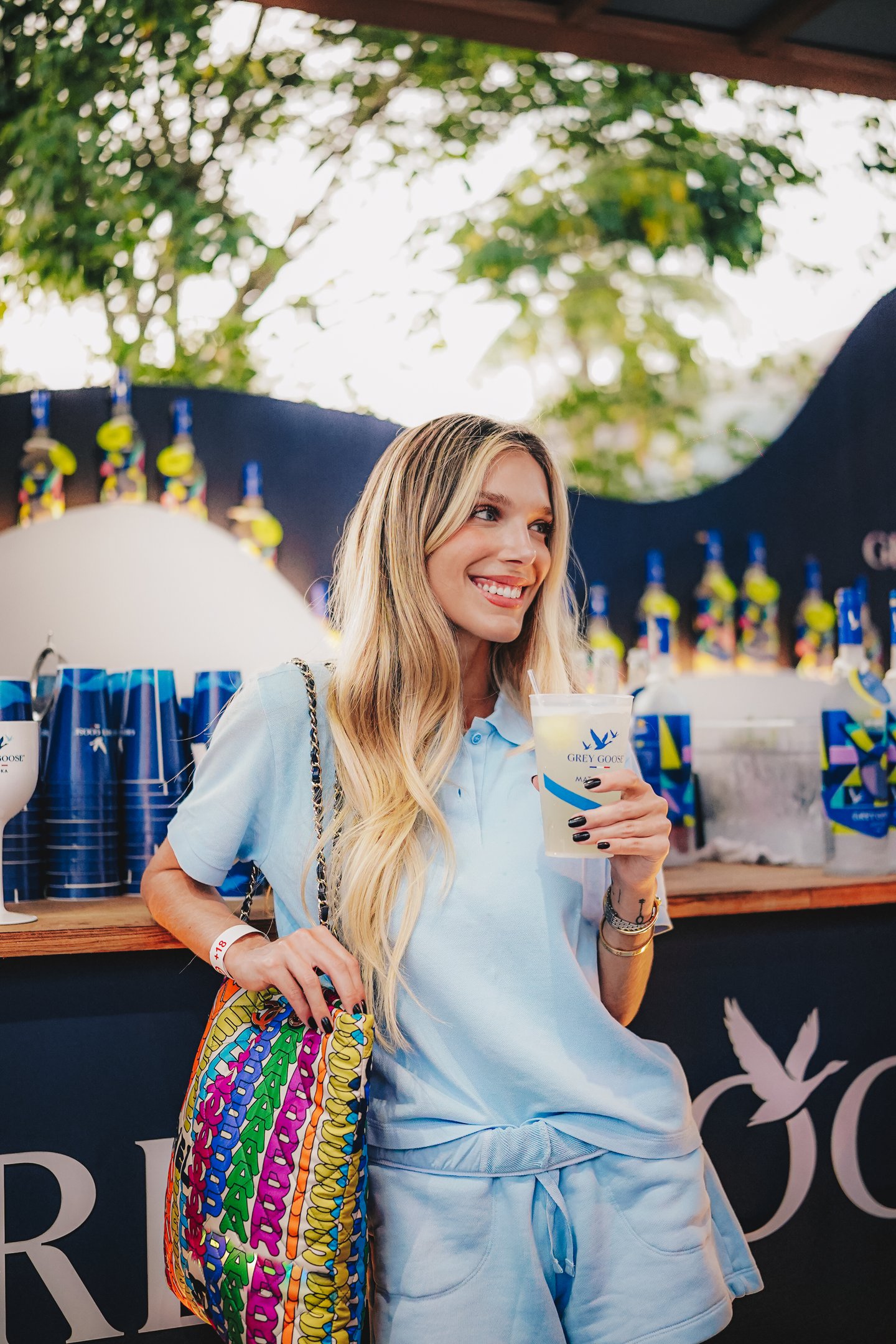 Mulher sorrindo usando conjunto azul bebe  em frente a logo da Grey Goose. Ambiente descontraído no contexto do Rio Open.