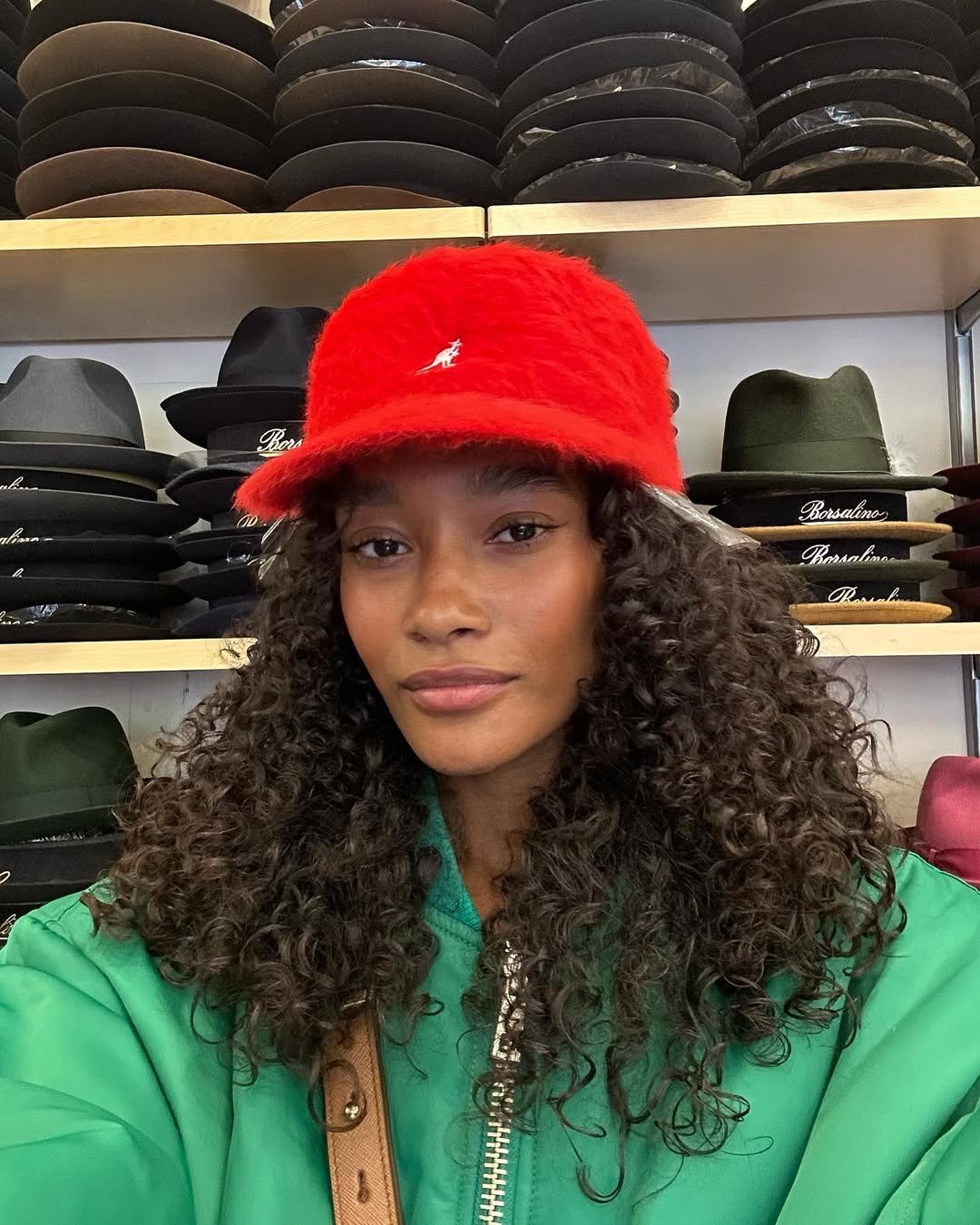 Pessoa com cabelo cacheado veste boné vermelho. Em fundo, prateleira com chapéus variados. Guia Do Cabelo Cacheado