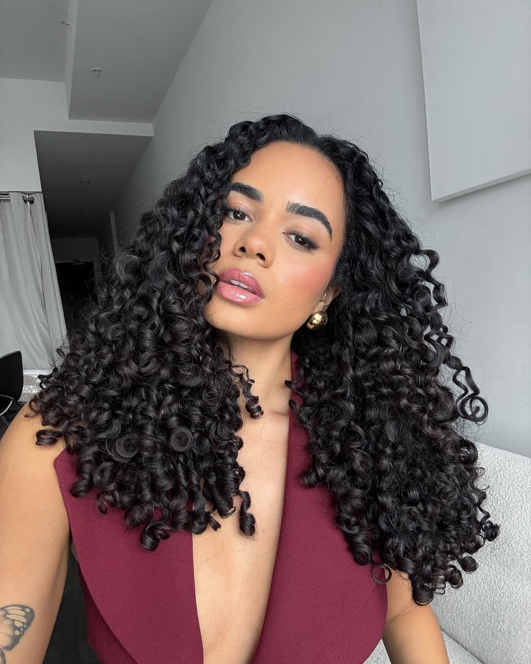 Mulher com cabelos cacheados definidos, usando top vinho. Os cachos ilustram um estilo do Guia do cabelo cacheado.