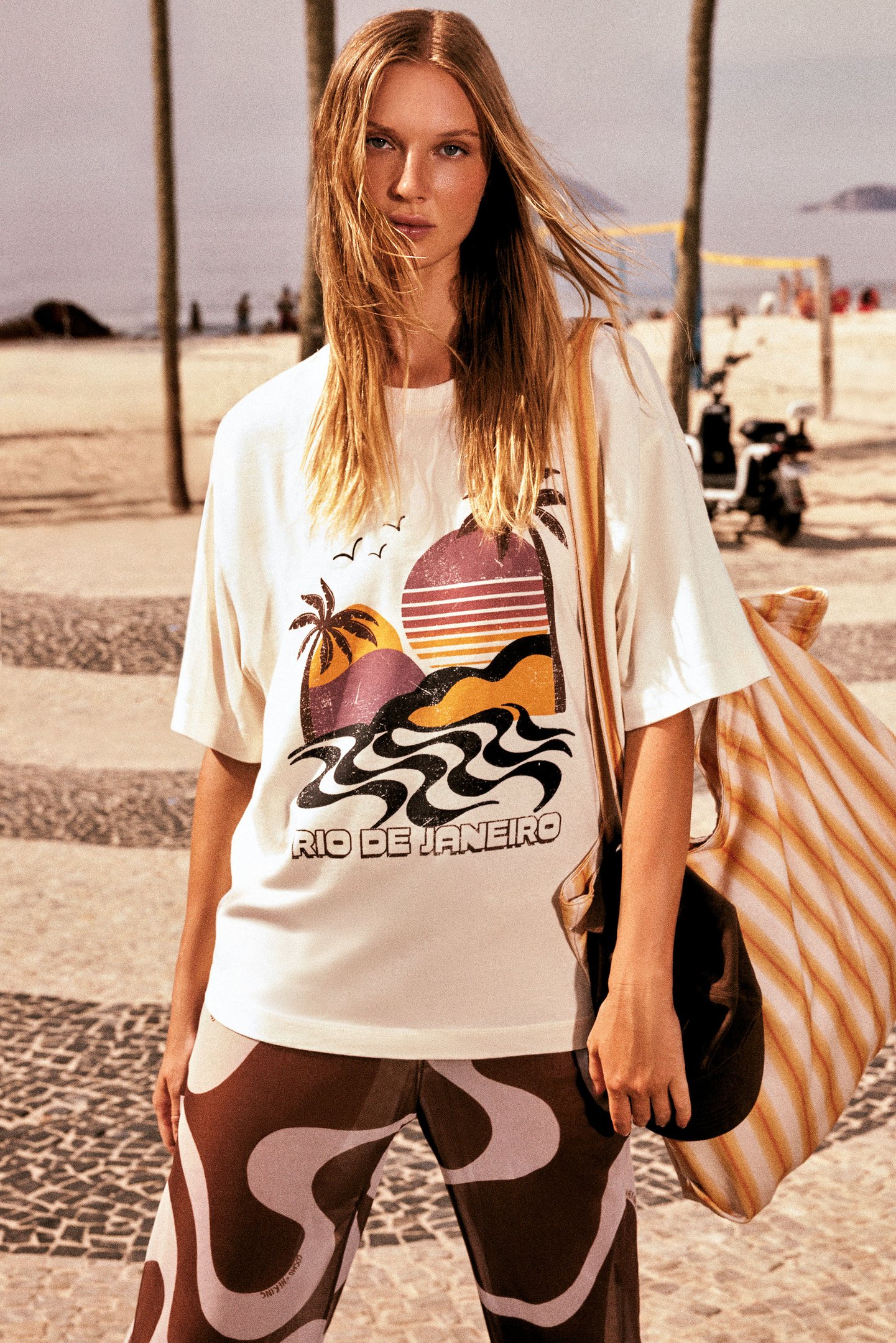 Pessoa vestindo camiseta com estampa do Rio de Janeiro, segurando bolsa listrada em praia.