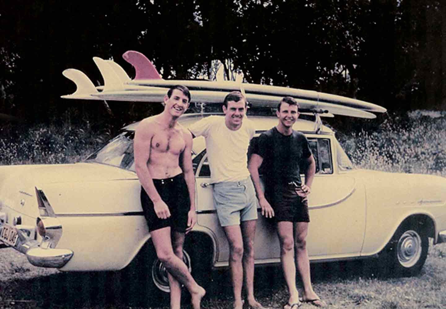 Três surfistas posam ao lado de um carro com pranchas no teto, remetendo ao início da história da UGG.
