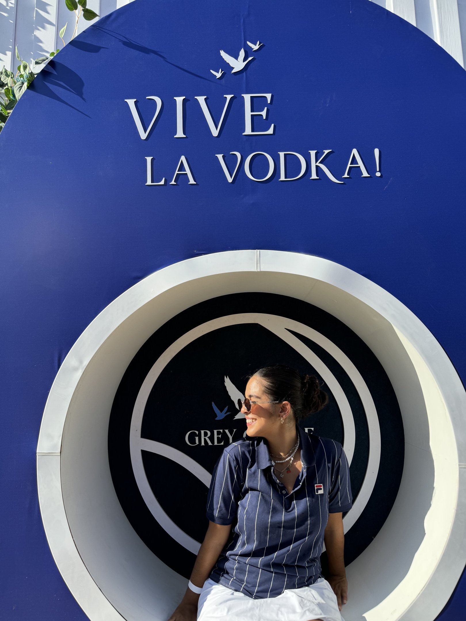 Pessoa sorrindo sentada em frente a um logotipo de vodka com fundo azul no evento Rio Open 