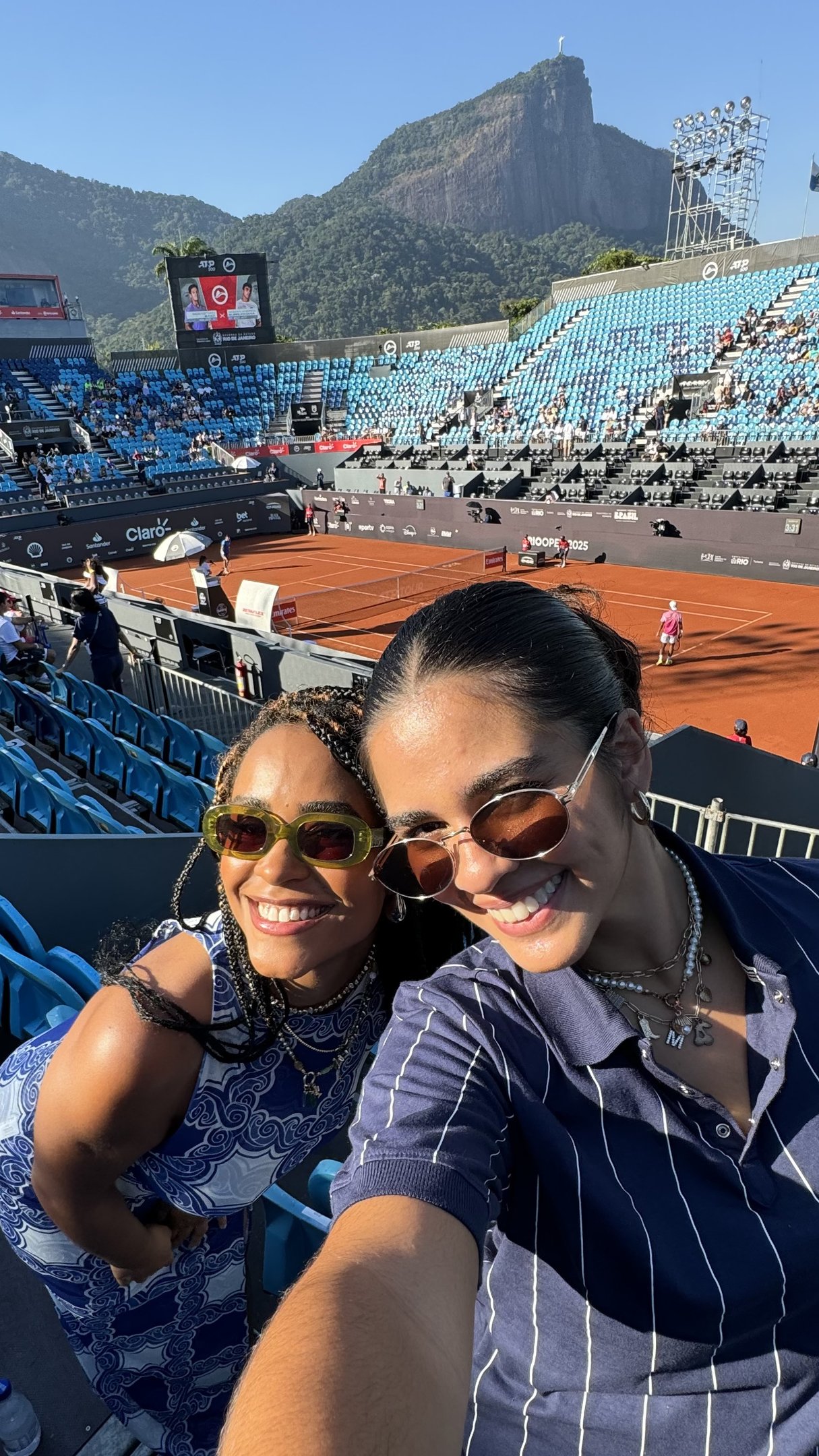 Pessoas sorrindo em uma arquibancada durante o torneio Rio Open, com montanha ao fundo.