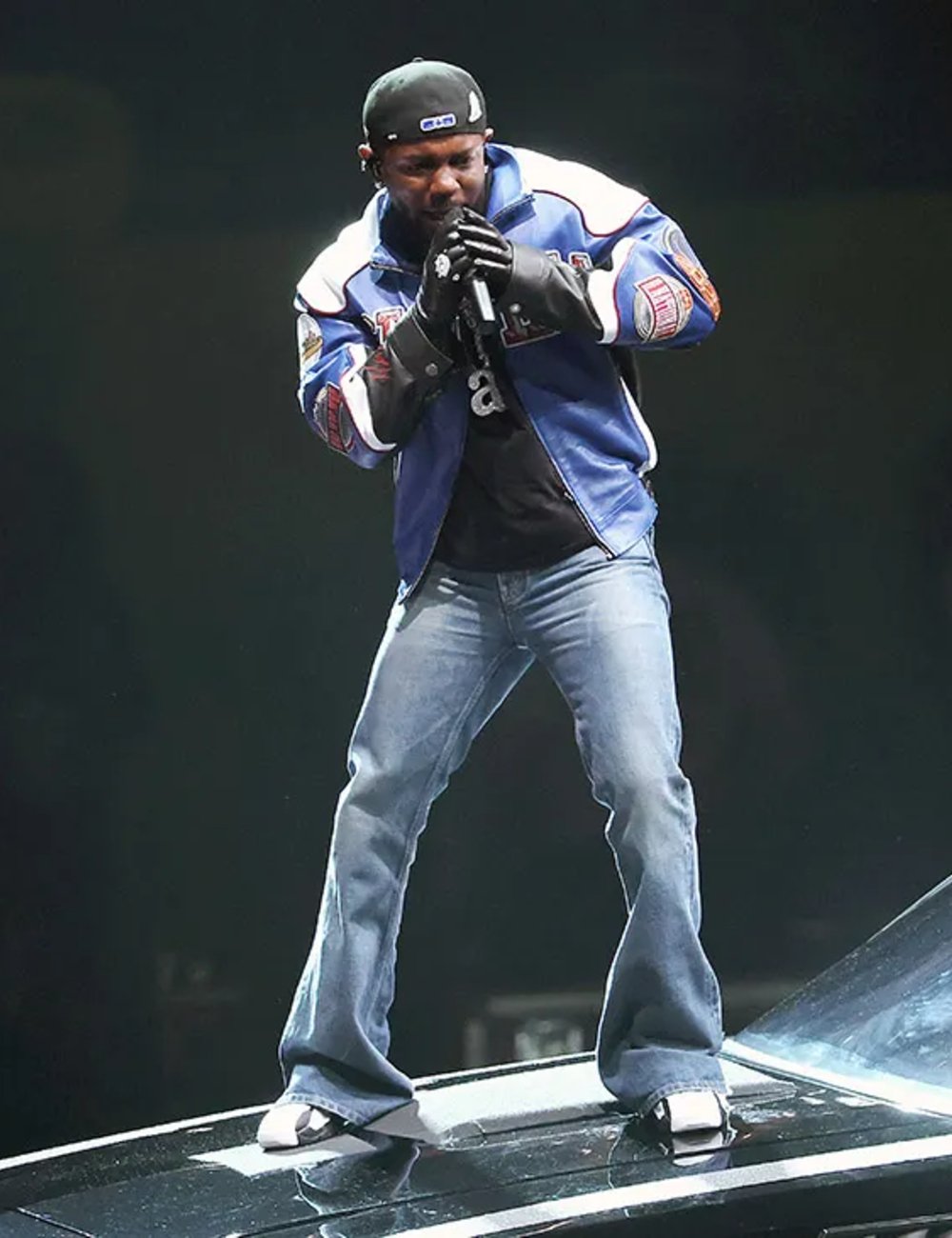Kendrick Lamar usando jaqueta azul e calça jeans atua no palco segurando um microfone.