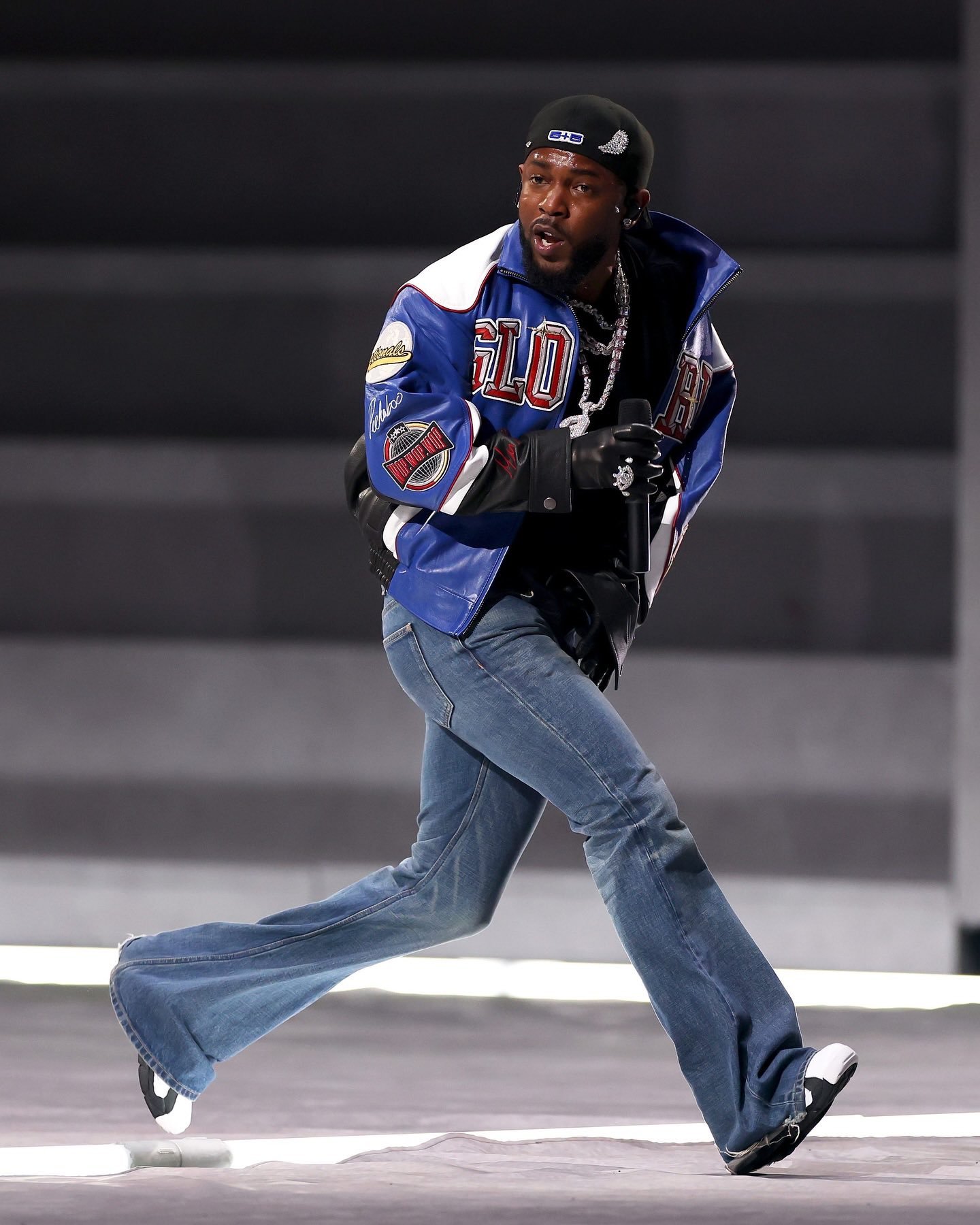 Kendrick Lamar está performando no palco, usando jaqueta azul e calça jeans.