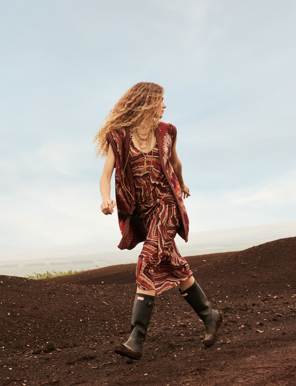 Modelo caminha sobre solo escuro em look com vestido estampado, colete e botas. Destaque para estilo e lançamentos de moda.