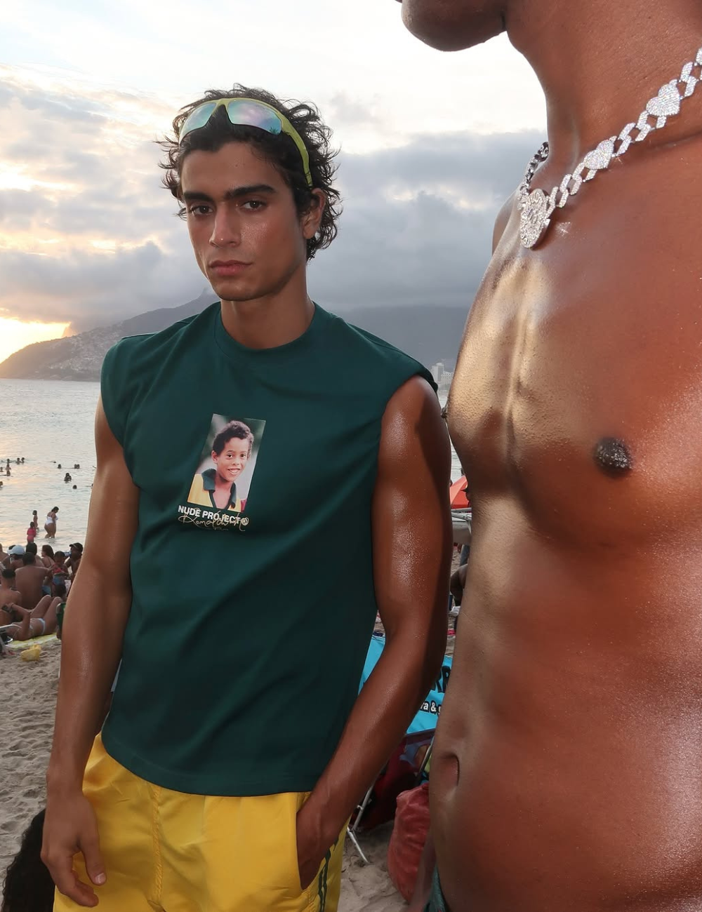 Modelo em praia usando camiseta estilosa, combinando com shorts amarelos. Acessório chama atenção.