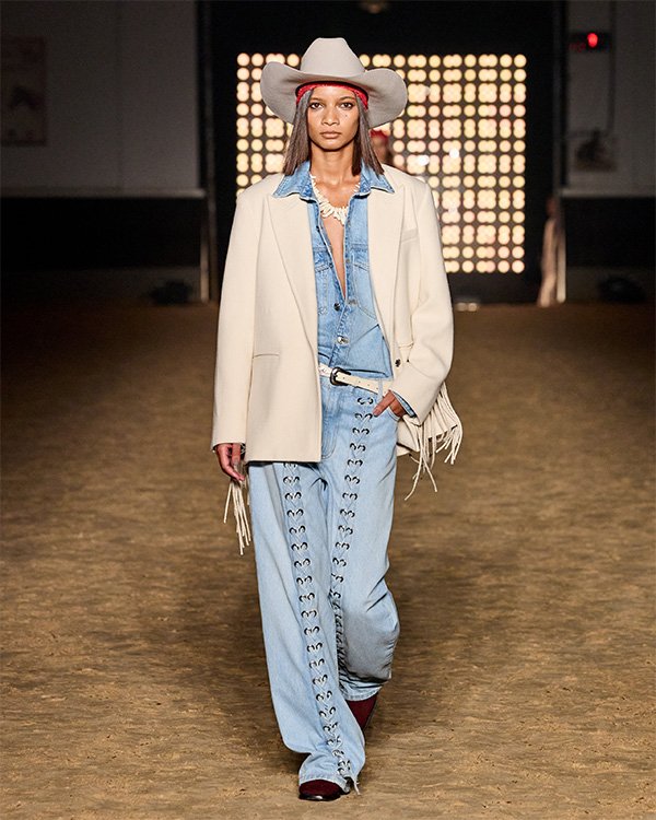 Desfile de moda com traje country, incluindo jaqueta branca e calça jeans, complementado por um chapéu de vaqueiro.