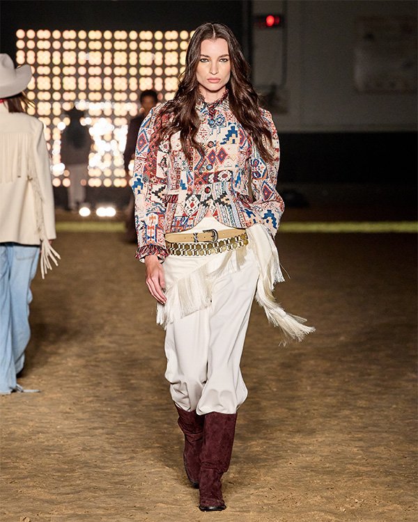 Modelo em desfile de moda veste blusa estampada tribal, calças brancas e botas marrons, em passarela iluminada.