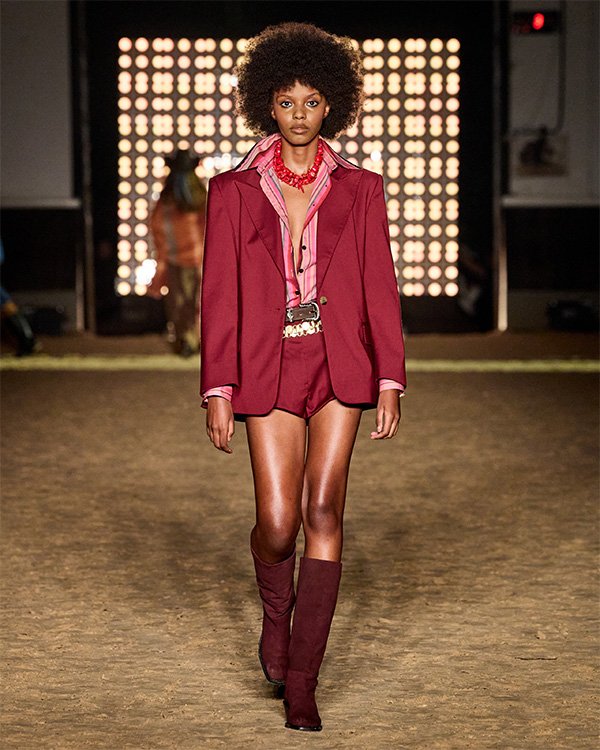 Desfile de moda com modelo em conjunto vermelho, composto por jaqueta, shorts e botas, usando colar vermelho.

