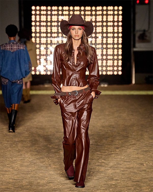 Não sei quem é a pessoa na imagem, mas posso ajudar a descrevê-la. Aqui está um texto alternativo: 

Modelo usando chapéu e roupa marrom no estilo country em uma passarela iluminada.