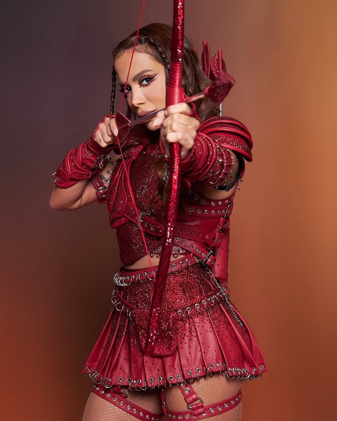 Anitta vestida em traje vermelho segurando arco e flecha.