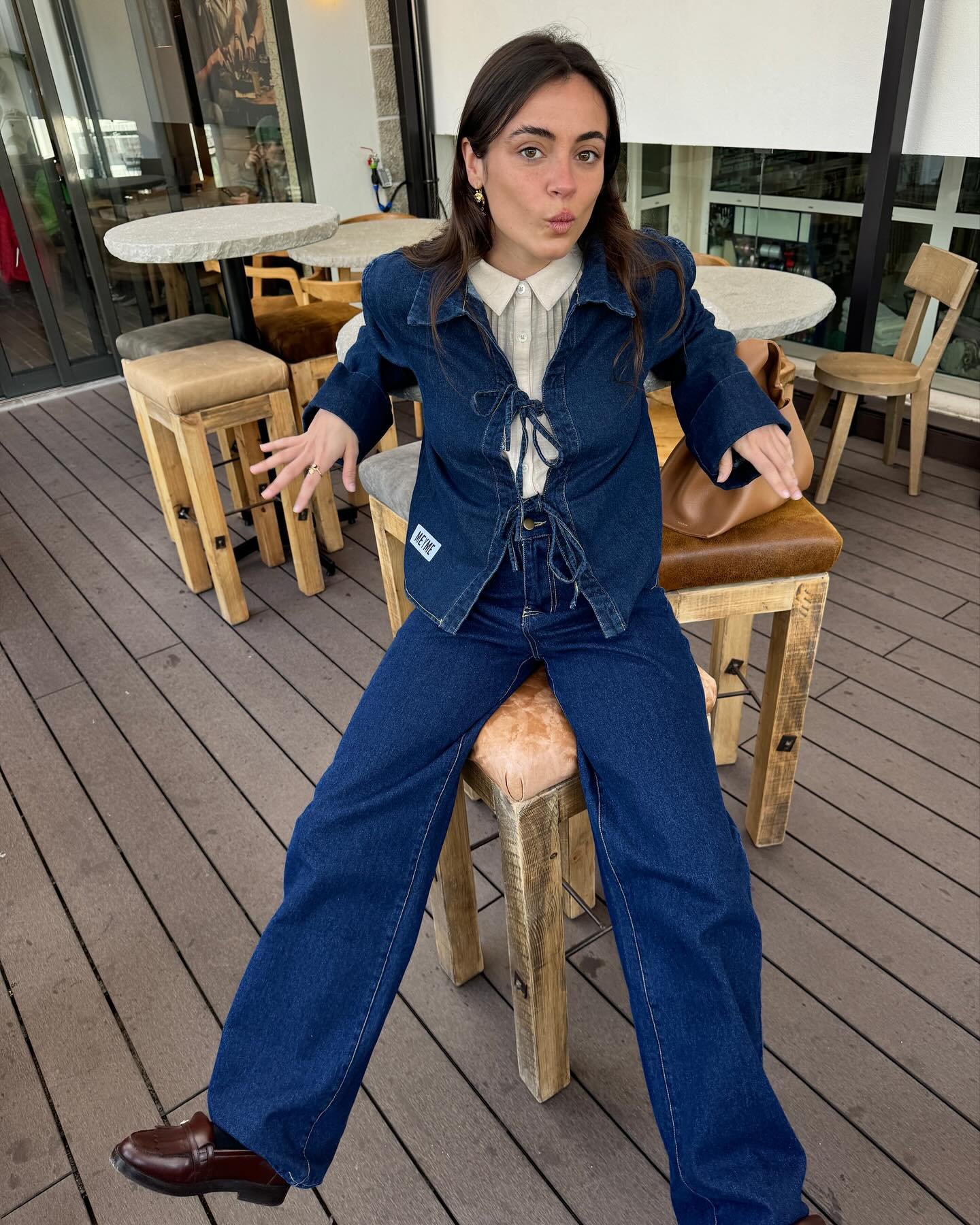 Pessoa vestindo conjunto jeans em ambiente com mesas e cadeiras de madeira, expressando-se de forma descontraída.
