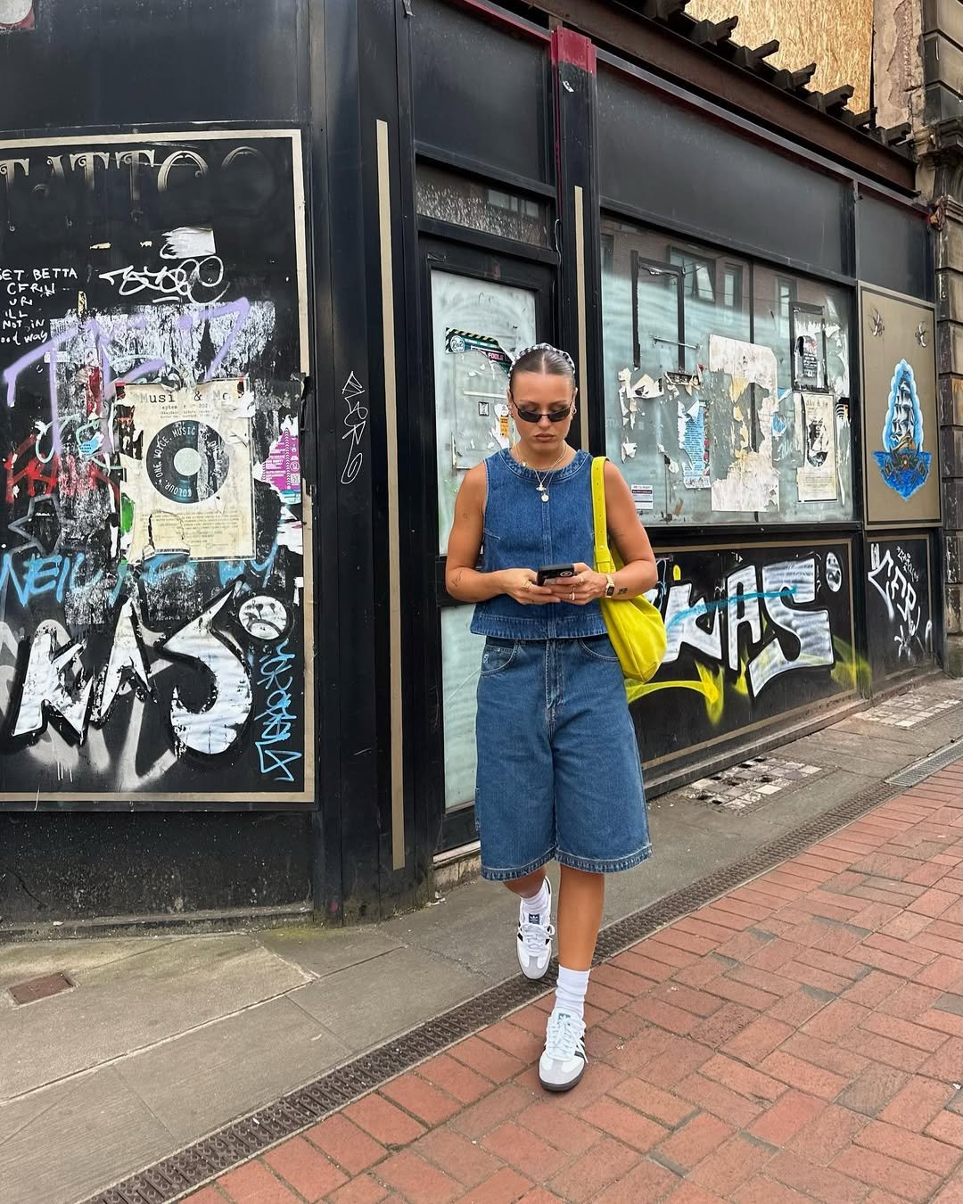 Pessoa vestindo jeans, composta por colete e bermuda, caminha na rua com grafites ao fundo, segurando celular e bolsa amarela.