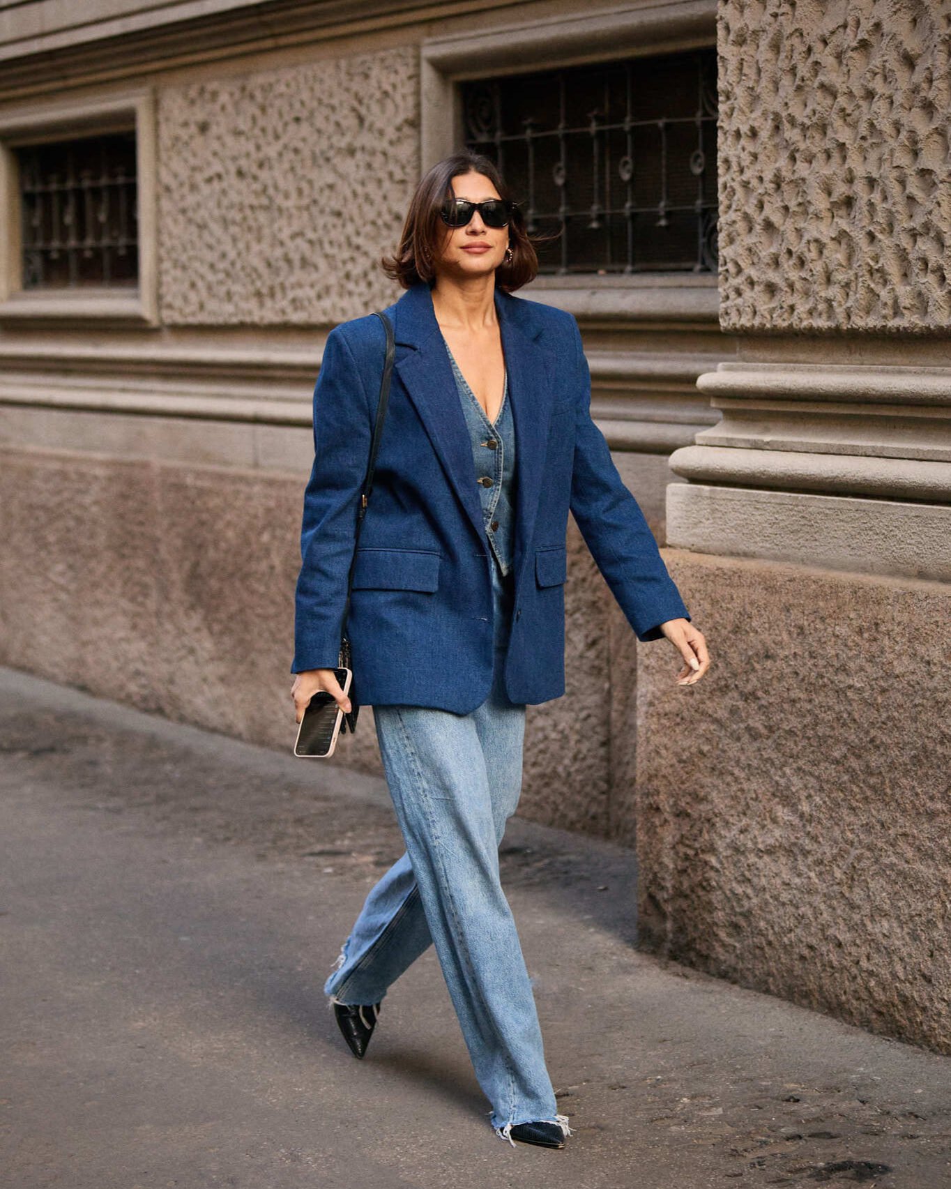 Pessoa caminhando na rua com roupa all jeans: blazer e calça jeans. Ela usa óculos escuros e carrega um celular.