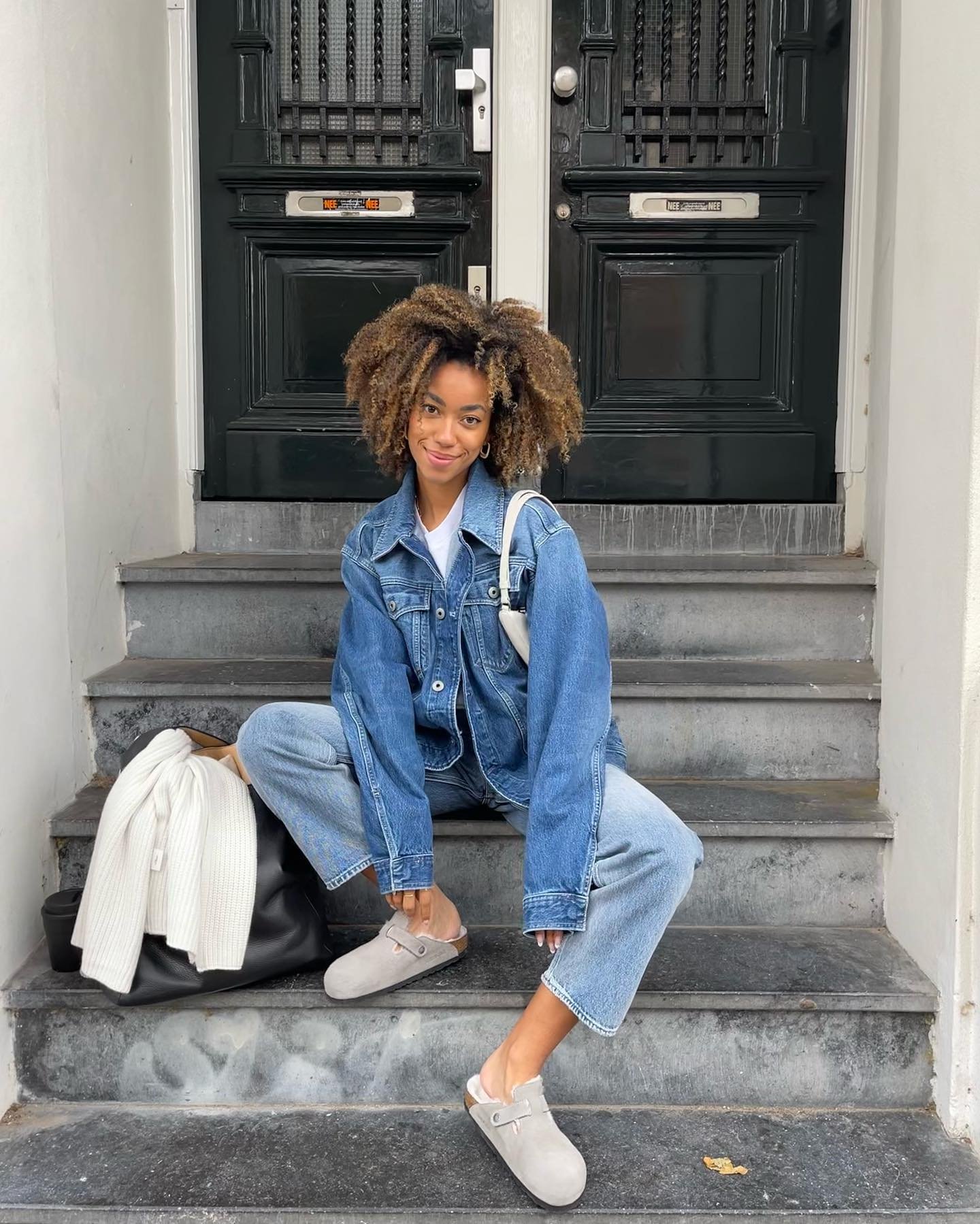 Pessoa sentada em escadas usando jaqueta e calça jeans, look all jeans, baguete branca no colo e bolsa preta ao lado.