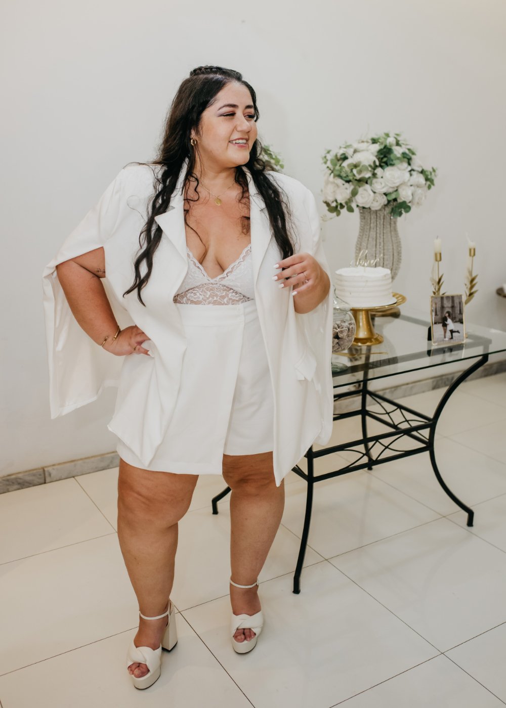 looks de noiva plus size: Layla em traje branco elegante, com flores ao fundo e decoração de casamento.