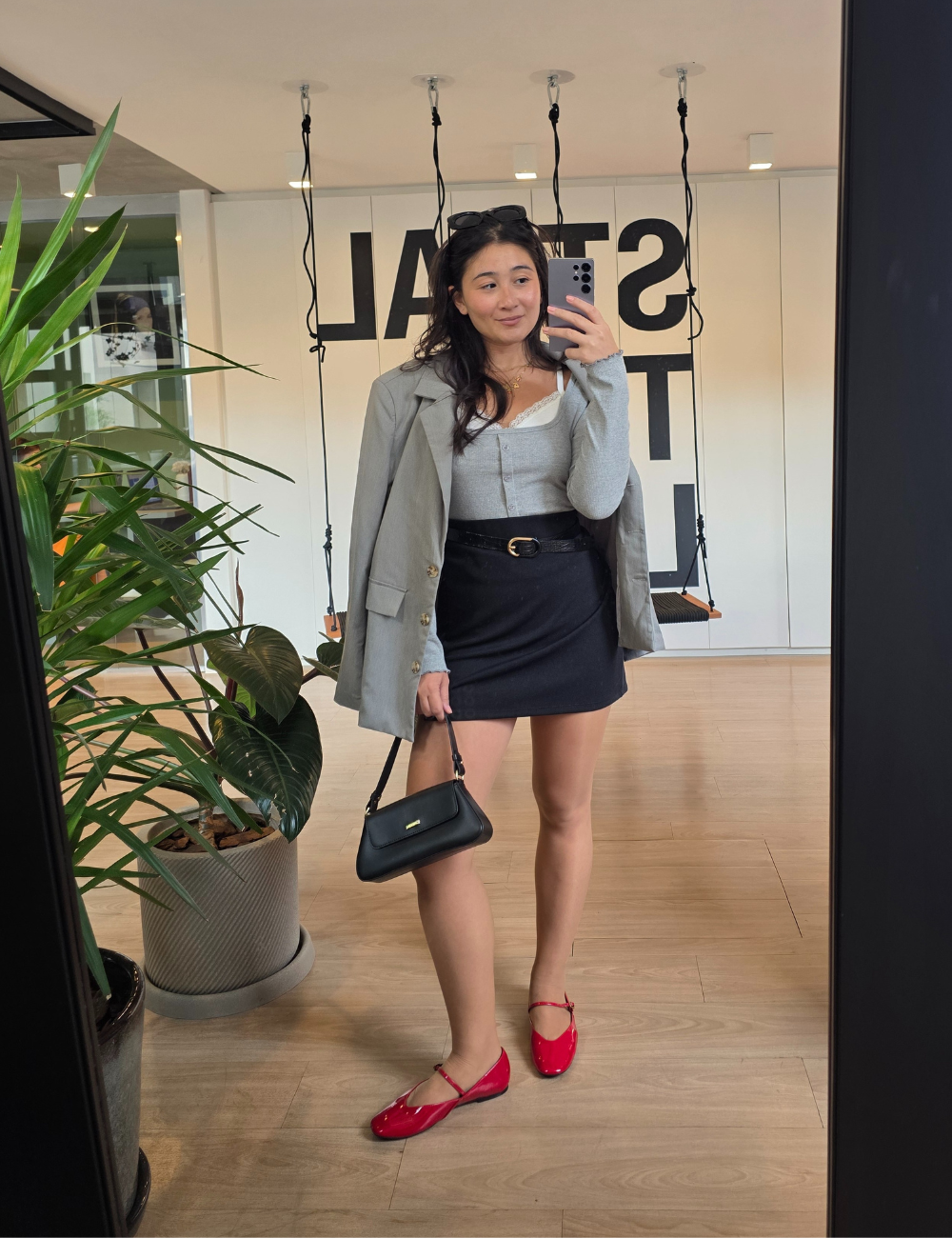 Mulher com blazer cinza, saia preta e sapatilhas vermelhas tira selfie em ambiente moderno com plantas.