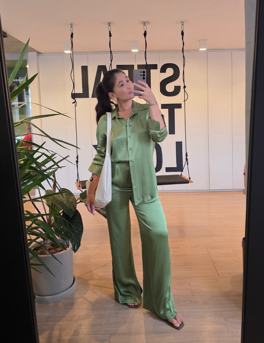 Mulher de roupa verde tirando selfie no espelho, segurando bolsa branca, com plantas e balanço atrás. Sugestão de look do ChatGPT.