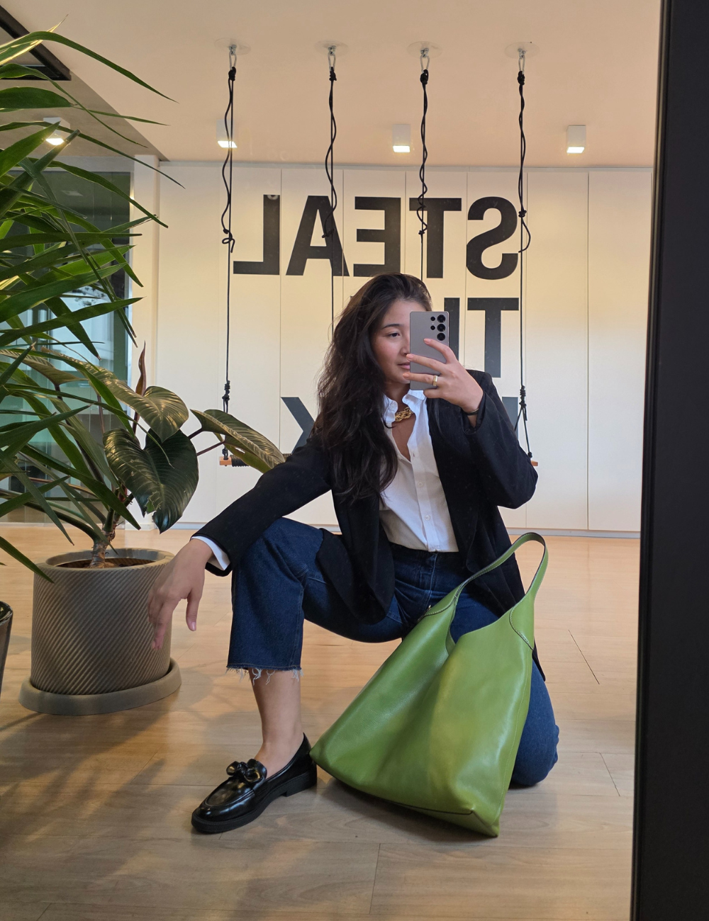 Pessoa ajoelhada em frente a um espelho, segurando um celular. Ela veste blazer preto, jeans rasgado e sapato preto, com bolsa verde.