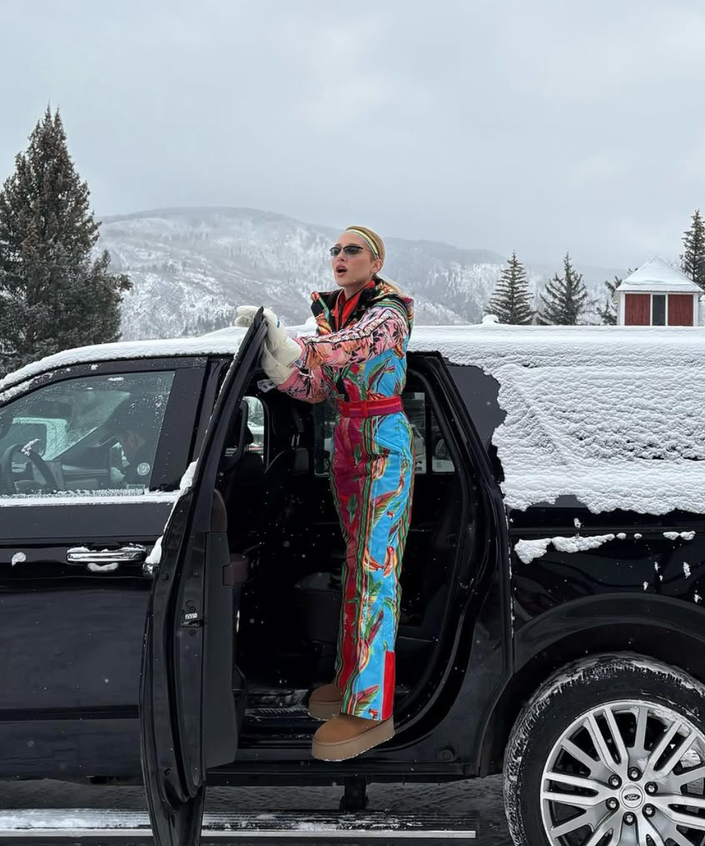Pessoa em macacão colorido de esqui ao lado de SUV coberto de neve em cenário montanhoso. Looks estilosos para esquiar.