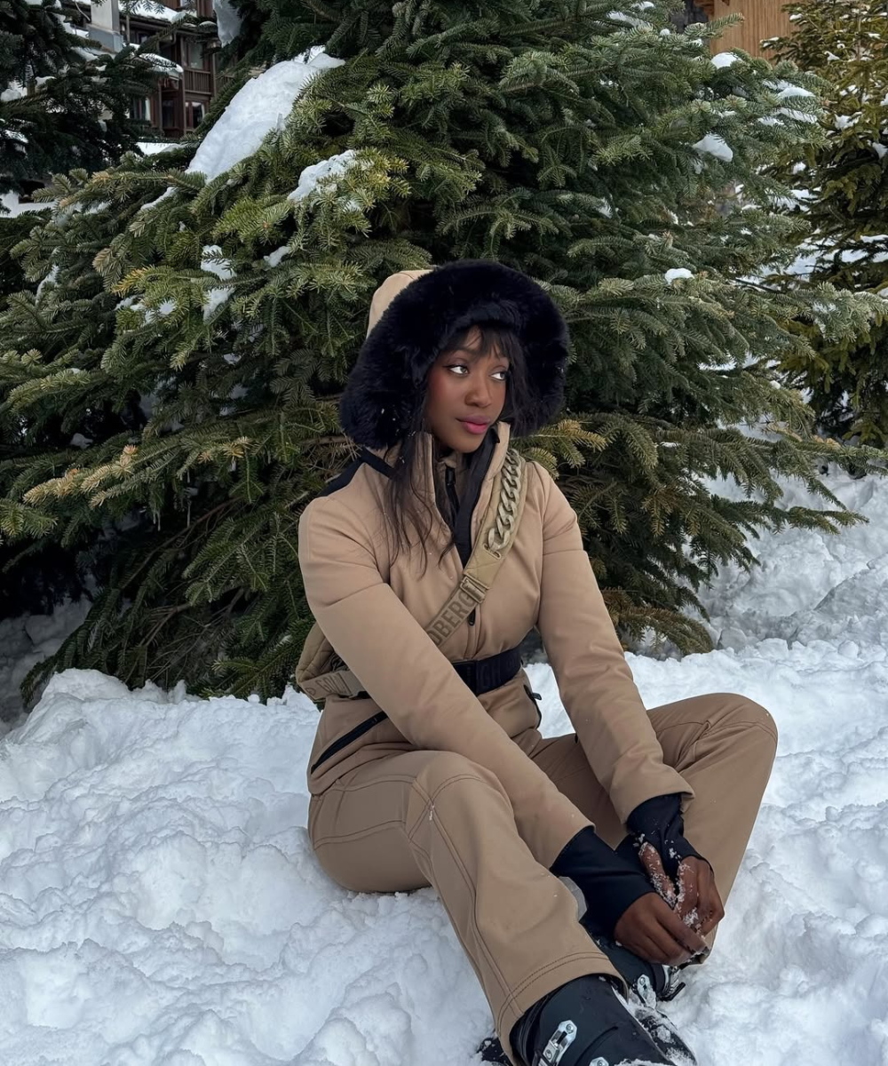 Pessoa sentada na neve em frente a pinheiros, vestindo roupa de esqui bege. Looks estilosos para esquiar.